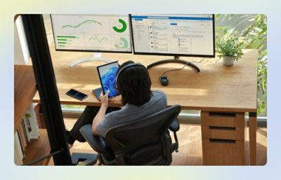Person working at a desk using a tablet, with two large monitors displaying charts and a dashboard.