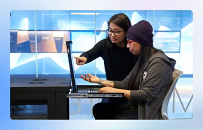 Two people at a computer: one seated in a beanie watching the screen, the other standing, pointing and explaining with focus.