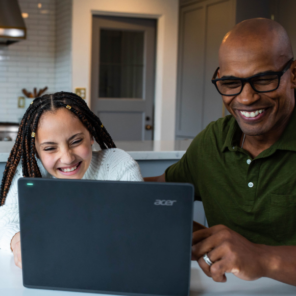 A father and daughter sitting together at a kitchen table at home looking at the screen of a laptop and smiling