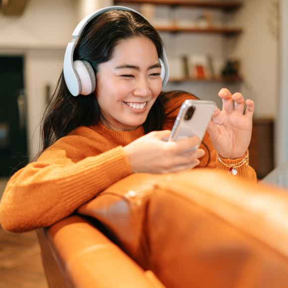 A young woman sitting on a sofa and listening to music with a wireless blue tooth headphones connected to her smart phone in her living room at home