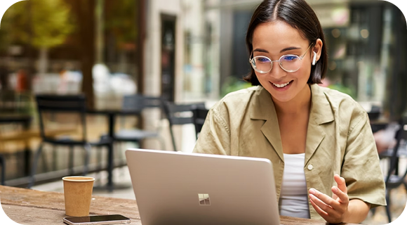 A person wearing glasses smiles and works on a laptop at an outdoor cafe