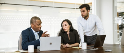Three people are gathered around a laptop in a modern office setting, engaged in discussion.