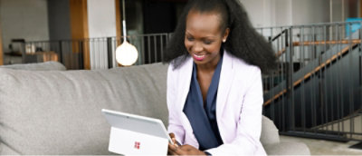 A woman in business attire sits on a gray couch, smiling while using a Microsoft Surface tablet in a modern office setting.