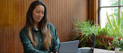 Person working on a laptop at a wooden table next to a window with a potted plant.