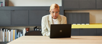 A person with blonde hair and glasses, wearing a light-colored blazer, sits at a desk working on a laptop 
