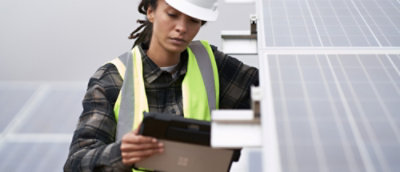 Ženski tehničar sa kacigom i zaštitnim prslukom pregleda solarni panel sa digitalnim uređajem.