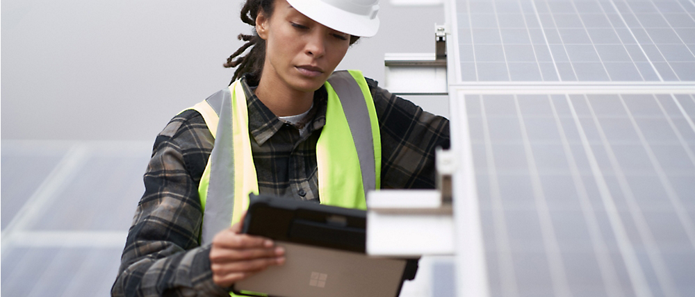 A female technician in a hard hat and safety vest inspects a solar panel with a digital device.