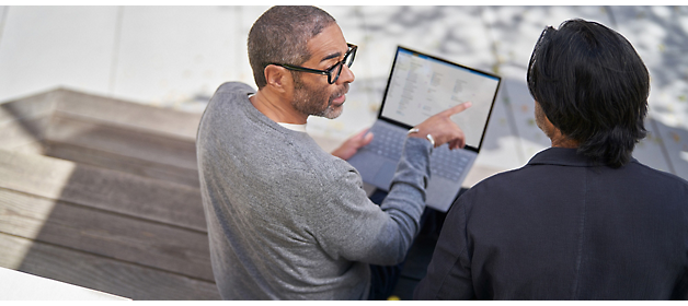 Dos hombres examinando la pantalla de un portátil juntos al aire libre, uno apuntando a la pantalla mientras participan en una discusión.