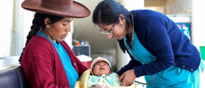 Zdravstveni radnik pregleda bebu koju drži žena u tradicionalnoj odeći na klinici.