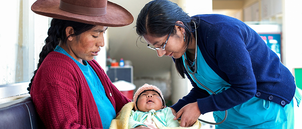 Bir sağlık çalışanı, bir klinikte geleneksel kıyafetli bir kadının kucağındaki bebeği muayene ediyor.