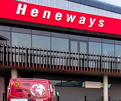 Exterior of a modern commercial building with a red “Heneways” sign across the upper фасade, glass windows, metal balcony railings, and a red van parked in front.