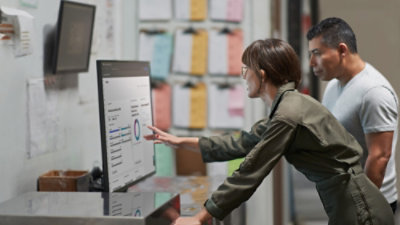 Two people standing indoors, reviewing a dashboard on a large monitor with charts and metrics displayed.