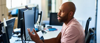 Person sitting at a desk speaking while looking at computer monitors.