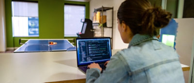 Person using a laptop displaying code in an office with a ping‑pong table in the background.