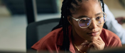  Person with braided hair wearing glasses and a rust‑colored shirt, seated at a desk.