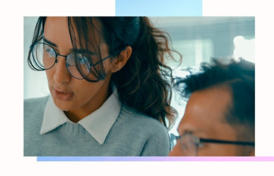 Two individuals in a workspace, with one wearing glasses and a collared sweater