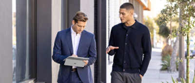 A man in a suit holding a tablet and looking at another man on a sidewalk.