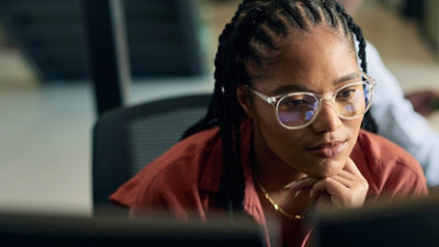 A person with braided hair sits at a desk, looking at computer monitors in an office setting.