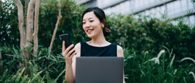 A woman holding a laptop and smiling while looking at the mobile