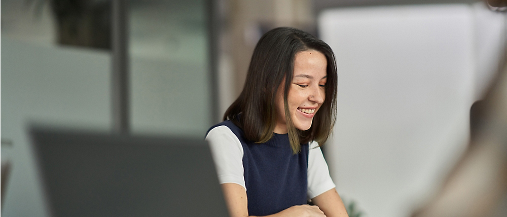 Une femme asiatique souriante, vêtue d’un haut bleu et blanc, assise à un bureau, en interaction avec un collègue invisible.