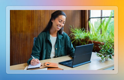 Person seated at a table using a laptop and writing notes in a plant‑filled workspace.