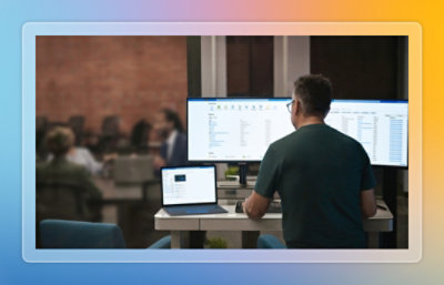 Person standing at an adjustable desk using a laptop and dual monitors in a collaborative office environment.