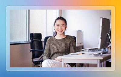 Person seated at an office desk with desktop computer, notebook, and papers in a bright workspace.