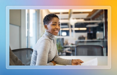 A person sits at a table holding papers in a glass-walled office meeting room with chairs and natural light.