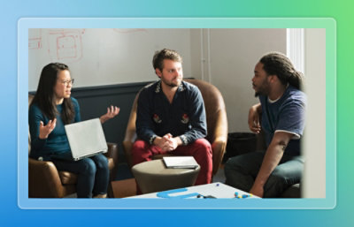 Three coworkers discuss ideas in a meeting, one using a laptop, others listening in a casual office setting.