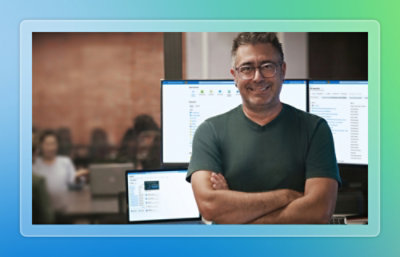 Person standing with arms crossed in an open office environment, with multiple computer monitors and coworkers visible in the background.