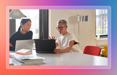 Two collaborators reviewing a laptop together across a table with notebooks nearby.