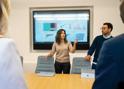 A presenter explains charts on a large screen during a team meeting in a conference room.