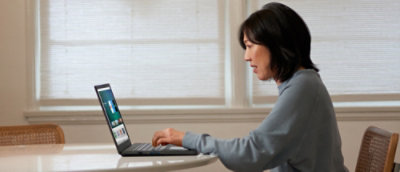 A person seated at a table using a laptop near a window in a bright indoor setting with natural light filtering through blinds.