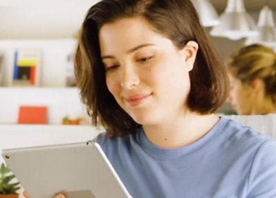 Person holding and using a tablet in a bright indoor workspace, with another person working in the background.