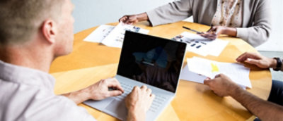 A close-up of a person typing on a laptop with a hand holding a pen nearby.