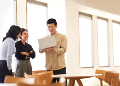 Three colleagues stand in a bright office discussing information on a laptop near a table and chairs.