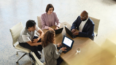 Four colleagues seated around a table reviewing content on laptops and a tablet in a meeting room.