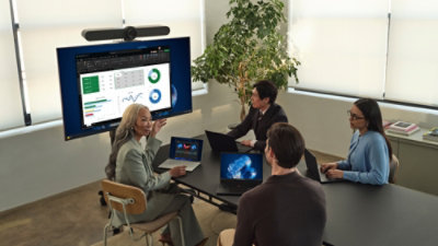 Team collaborating around a table during a meeting with laptops and a presentation screen.