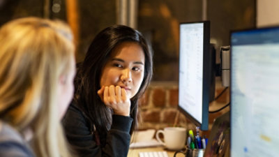 A person seated at a desk working on a computer in an office setting with dual monitors, a coffee mug, stationery items, and warm ambient lighting in the background.