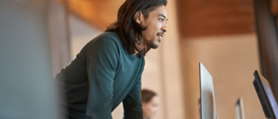 Person leaning toward desktop monitors while working in a modern office workspace.