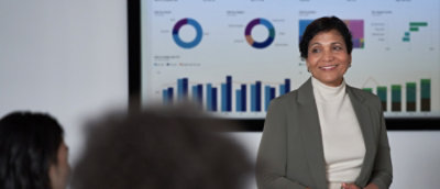 Presenter standing in front of a display showing charts and data summaries in a meeting room.