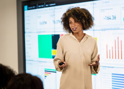 Presenter explaining analytics dashboards on a large display, holding a remote and gesturing during a data presentation.