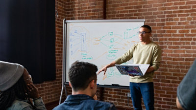 man explaining to participants while holding a laptop and standing in front of a whiteboard