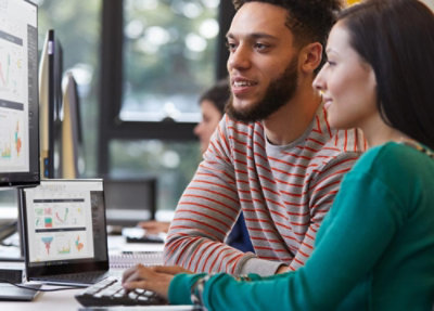 Two colleagues collaborating at a desk while reviewing data dashboards and charts displayed on a laptop and an external monitor in an office workspace.