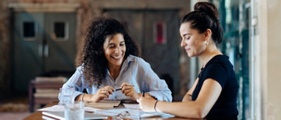 Two people seated at a table reviewing papers and notes together, with pens, a notebook, and a glass of water in a workspace.