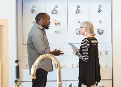 Two people discussing bathroom fixtures while standing in a showroom with various faucets and shower fittings displayed on the wall.