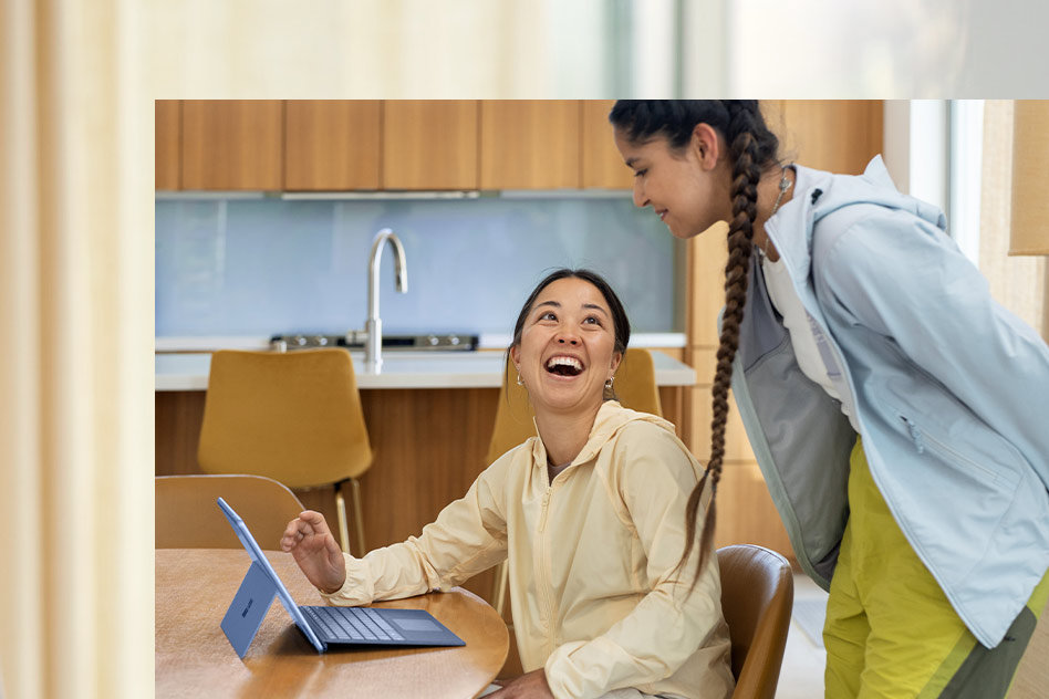 Två vänner vid ett köksbord skrattar åt en video på en Surface Pro 9-skärm.