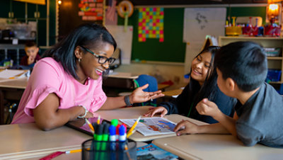 Eine Lehrerin lacht mit zwei Kindern in einem Klassenzimmer.