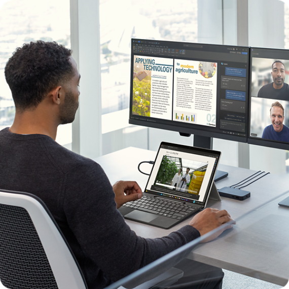 Man in a modern high-rise office sitting at a desk with multiple monitors connected to a Surface Pro featuring Live Captions.
