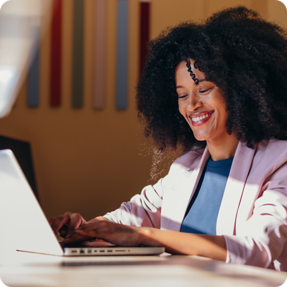 Cheerful African American businesswoman sitting at desk typing on laptop, working in modern office.